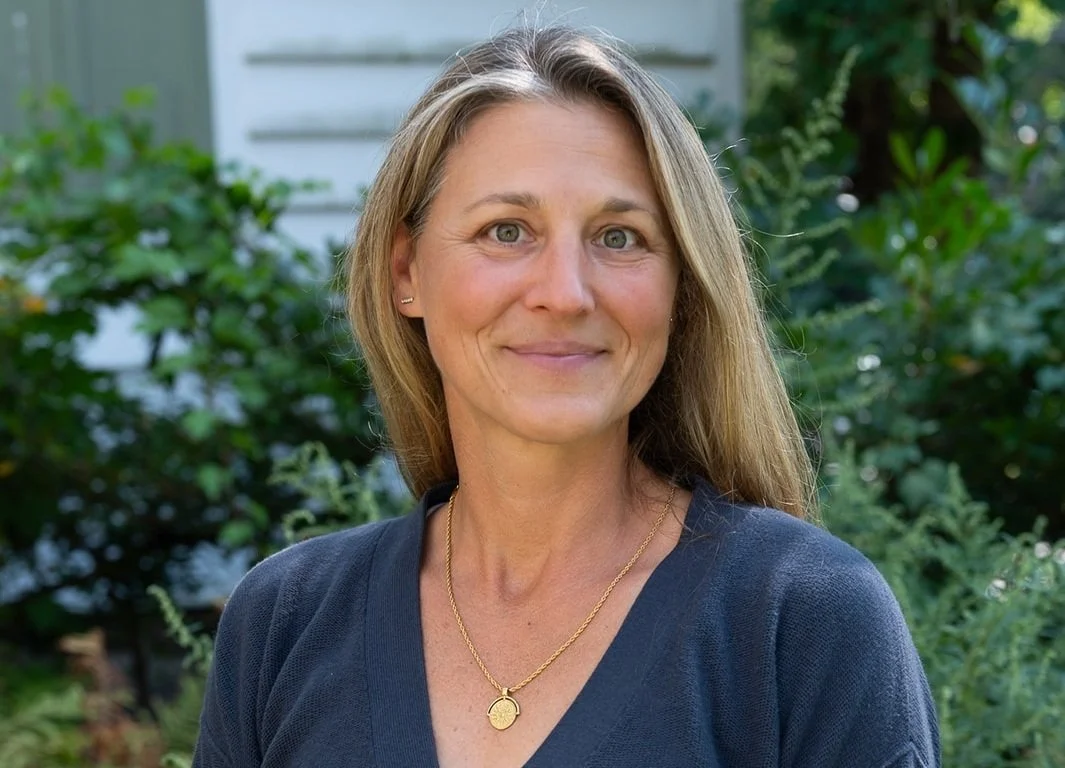 A woman with long blonde hair and blue eyes smiling outdoors, wearing a dark top and gold necklace, with green bushes and trees in the background.