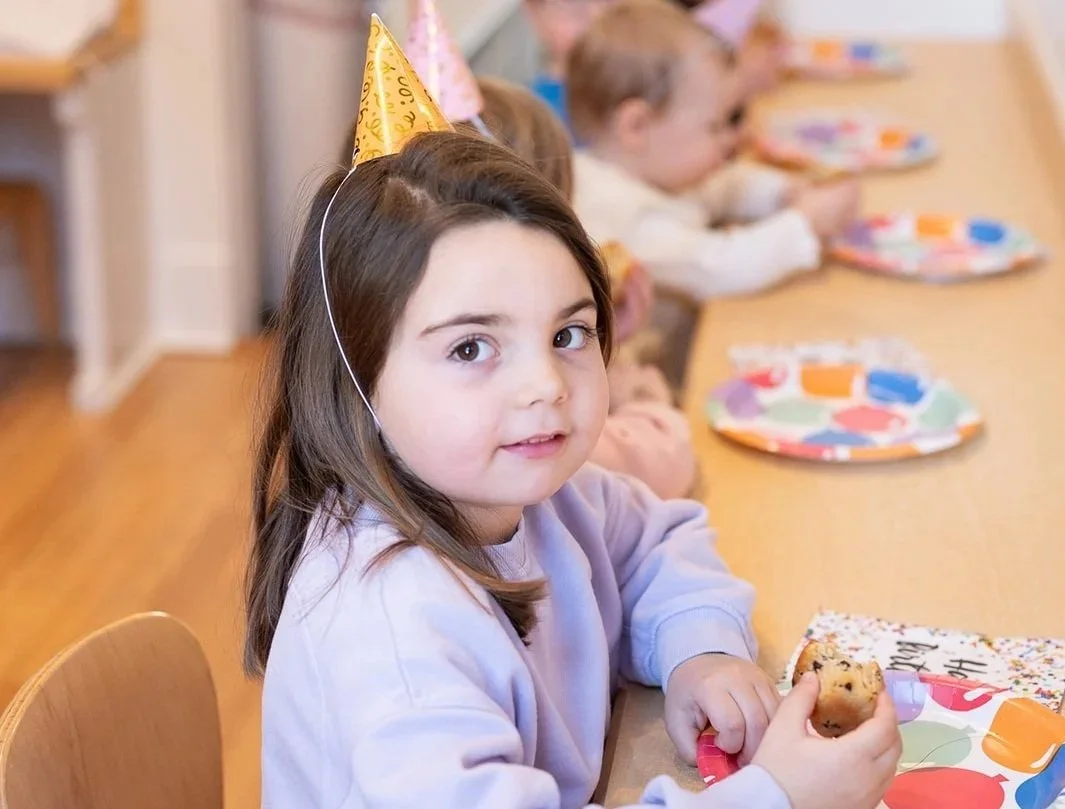 Young girl at a birthday party wearing a yellow party hat, sitting at a table with other children, holding a cookie, with colorful paper plates in front of her.