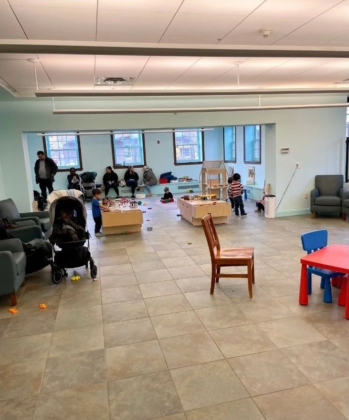Indoor playroom with children and adults, featuring toys, wooden playhouses, and seating, with large windows letting in natural light.