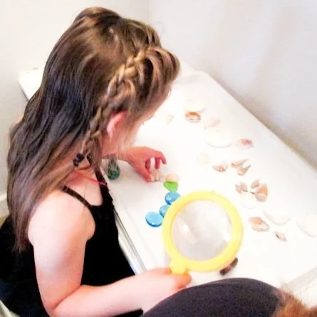 Young girl examining seashells on a table with a yellow magnifying glass.