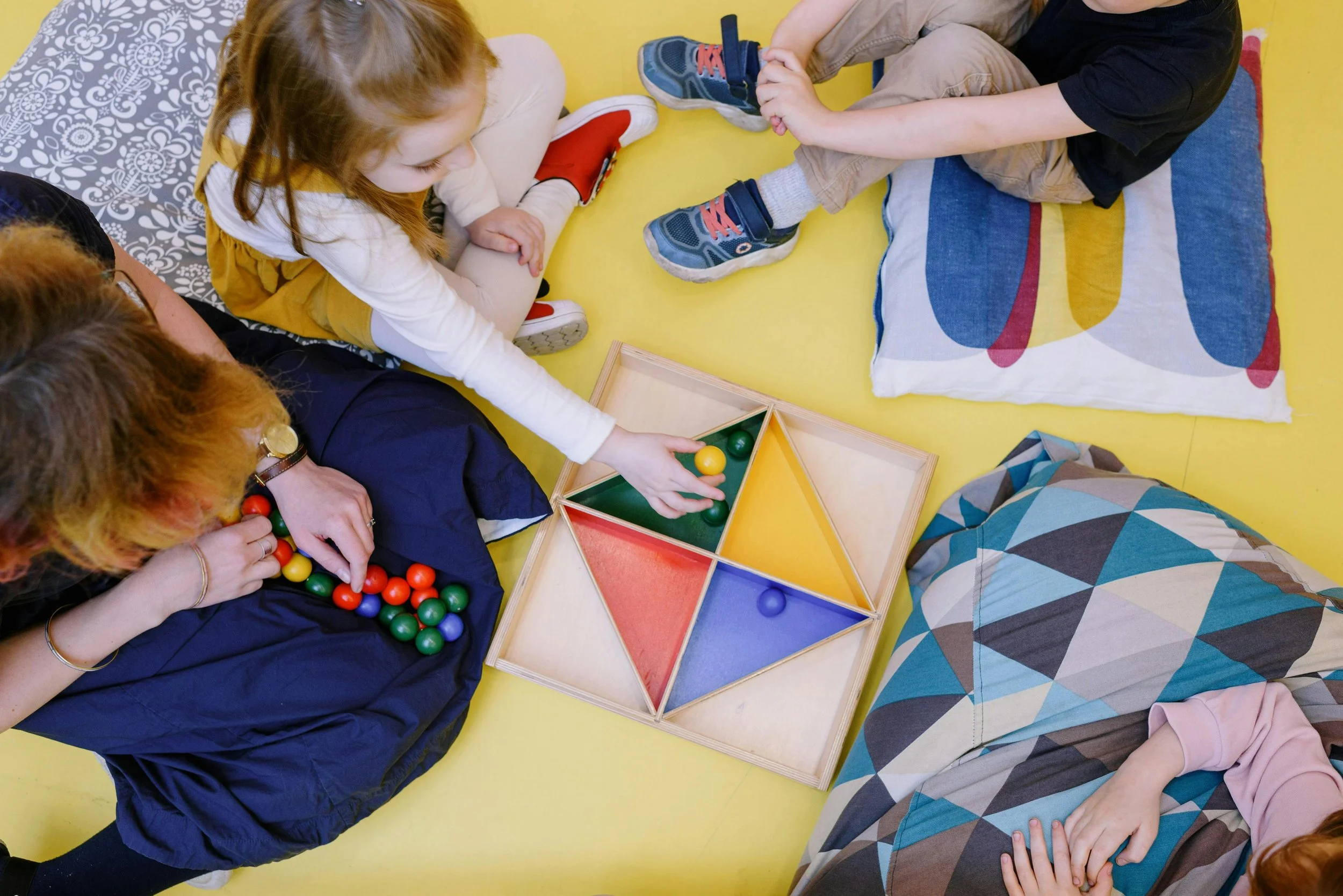 Children playing with colorful balls in a tray, sitting on a yellow floor, with pillows around them.