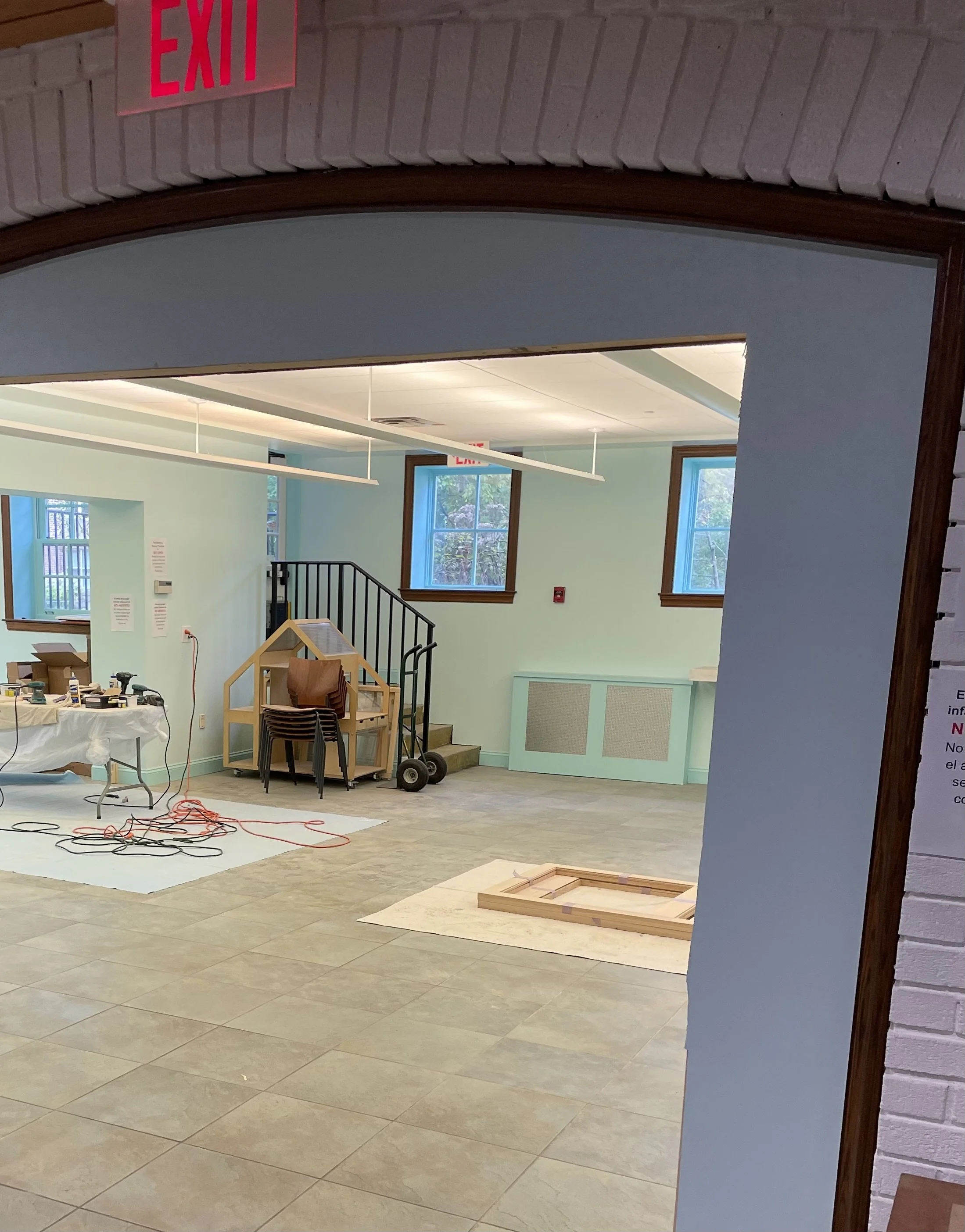 An indoor room under renovation with tile flooring, light green walls, two windows, and a staircase. There are construction materials, tools, and cords on the floor, and a partially assembled wooden frame on a rug. An 'EXIT' sign is visible on the ceiling.