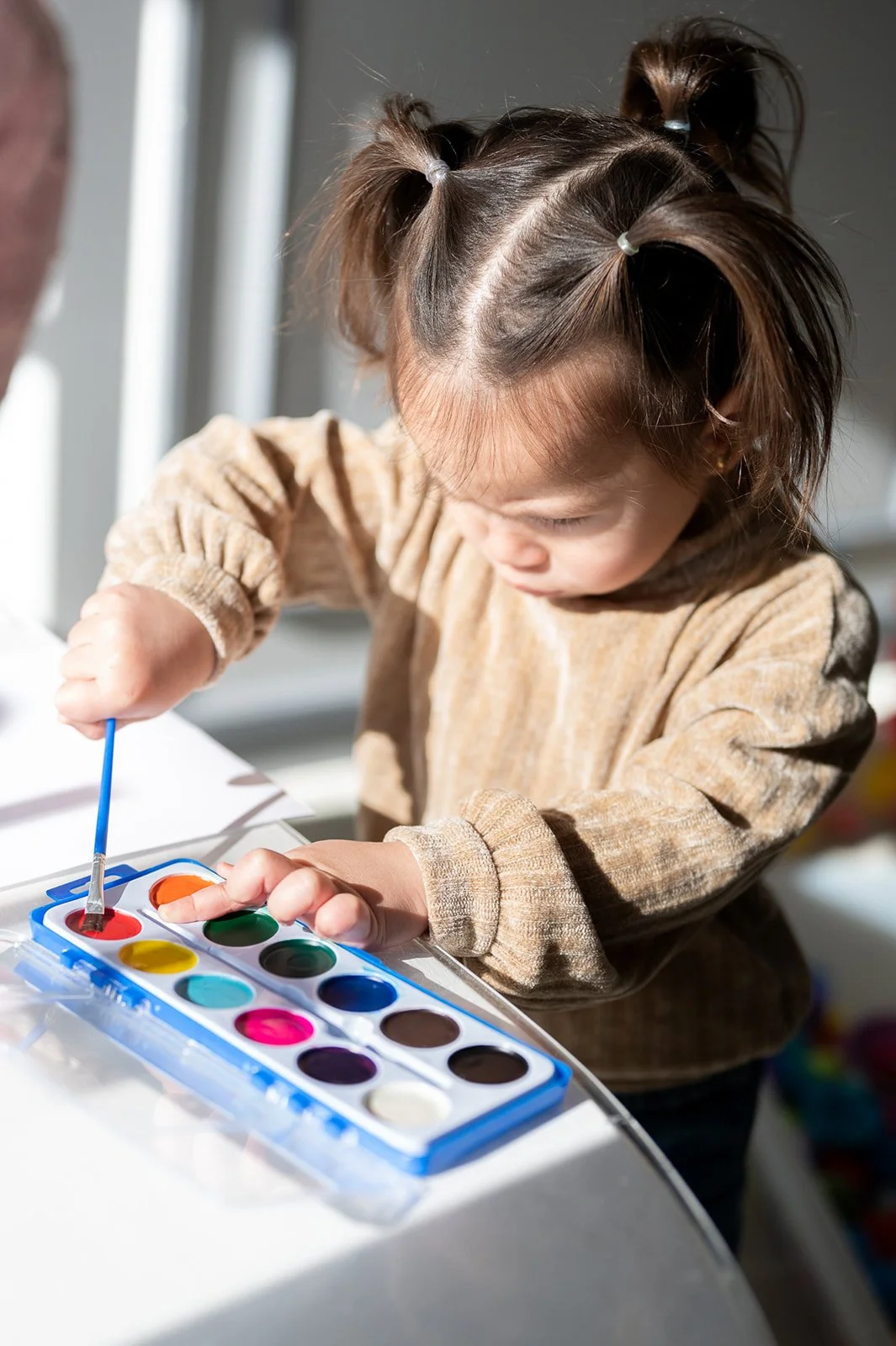 A young girl with brown hair styled in pigtails is painting with watercolors at a table.