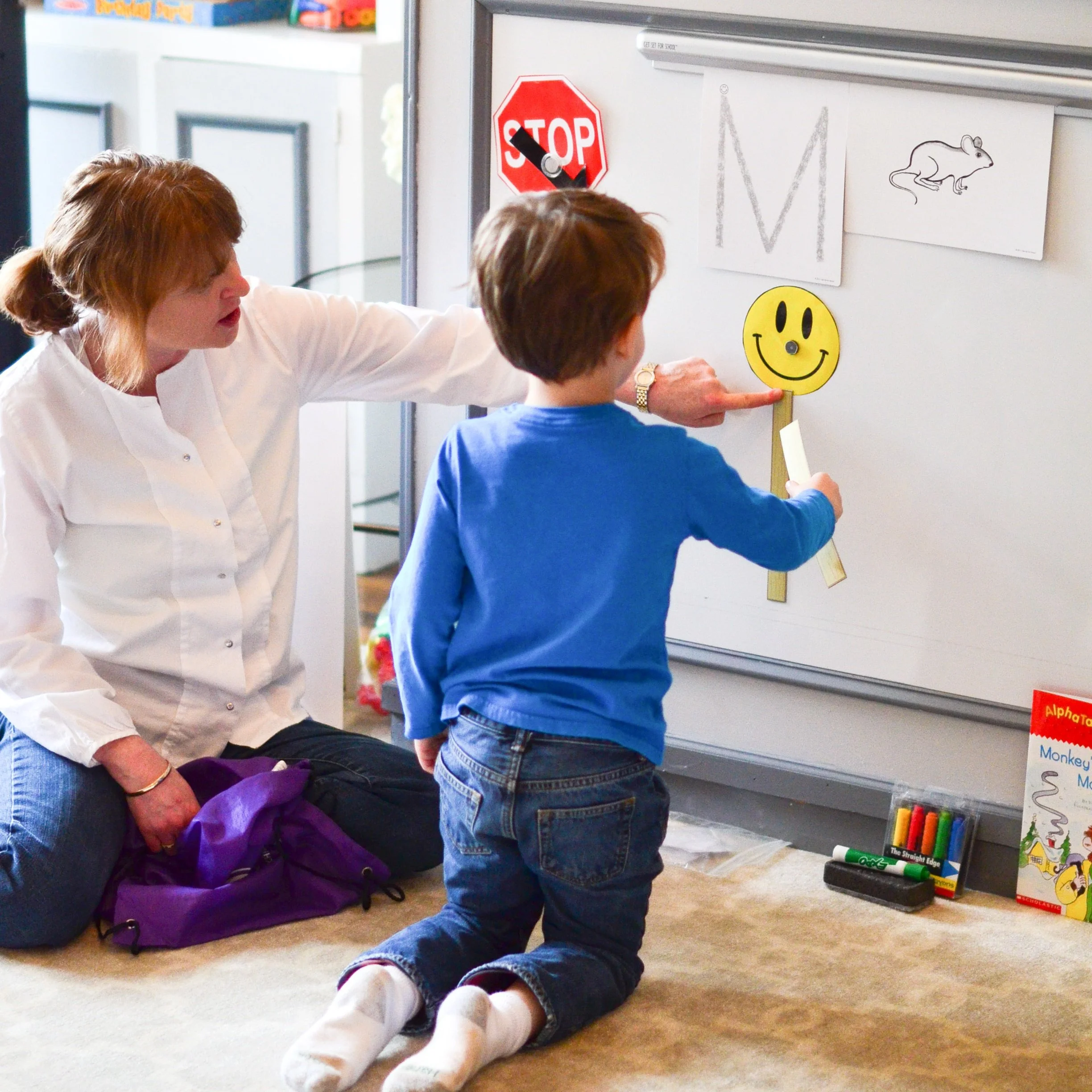 A young boy with brown hair kneels on the floor, pointing at a yellow smiley face sign held by a woman. The woman has red hair and is sitting next to him, wearing a white blouse. They are next to a whiteboard with various pictures and letters, including a stop sign and a letter M.
