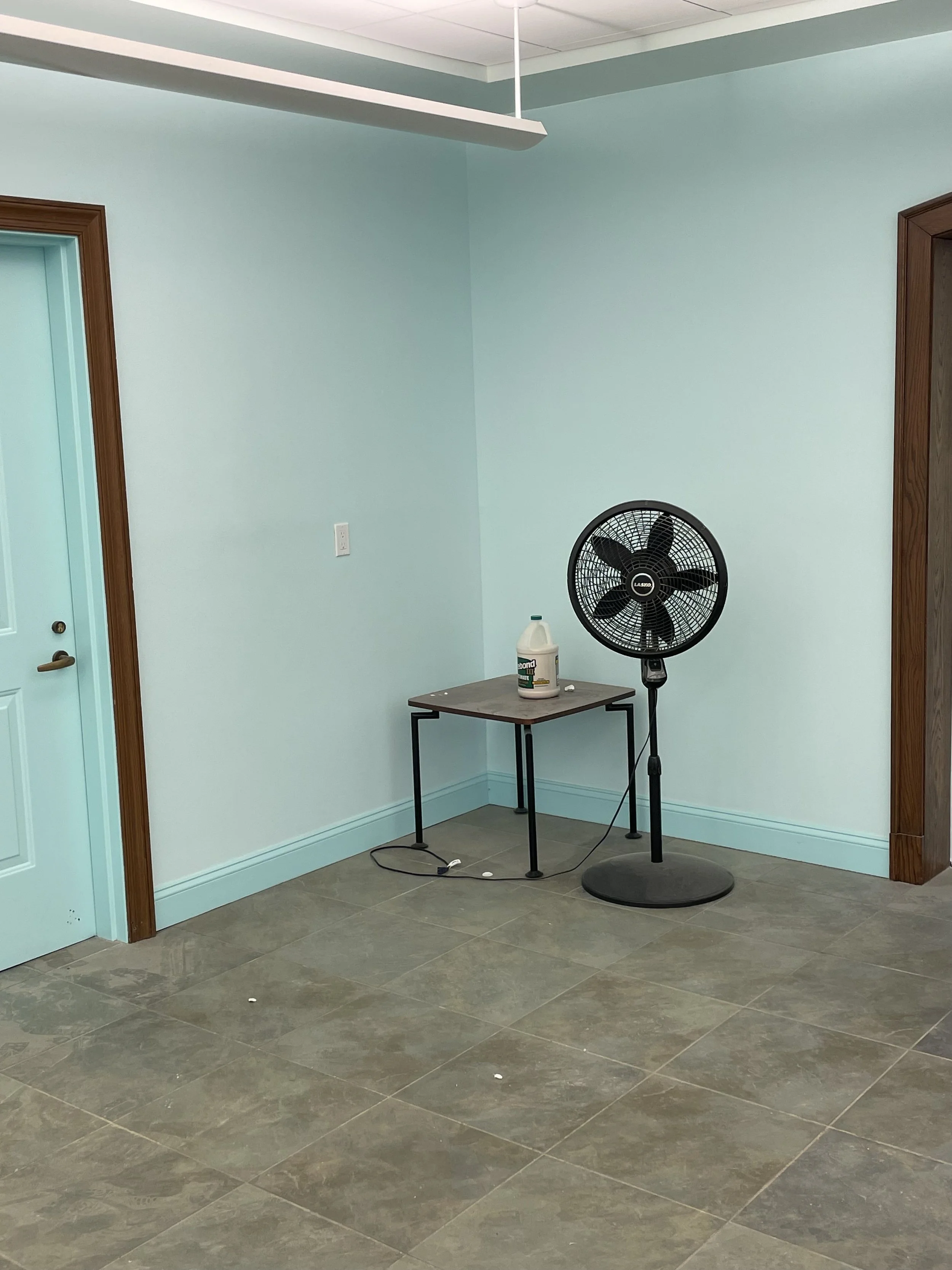 A room with light blue walls, brown door frames, a stand fan, a small table with a container of disinfectant, and tiled flooring.