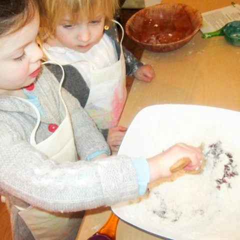Two children baking together, one mixing ingredients on a baking sheet while the other watches, in a kitchen with bowls and utensils.