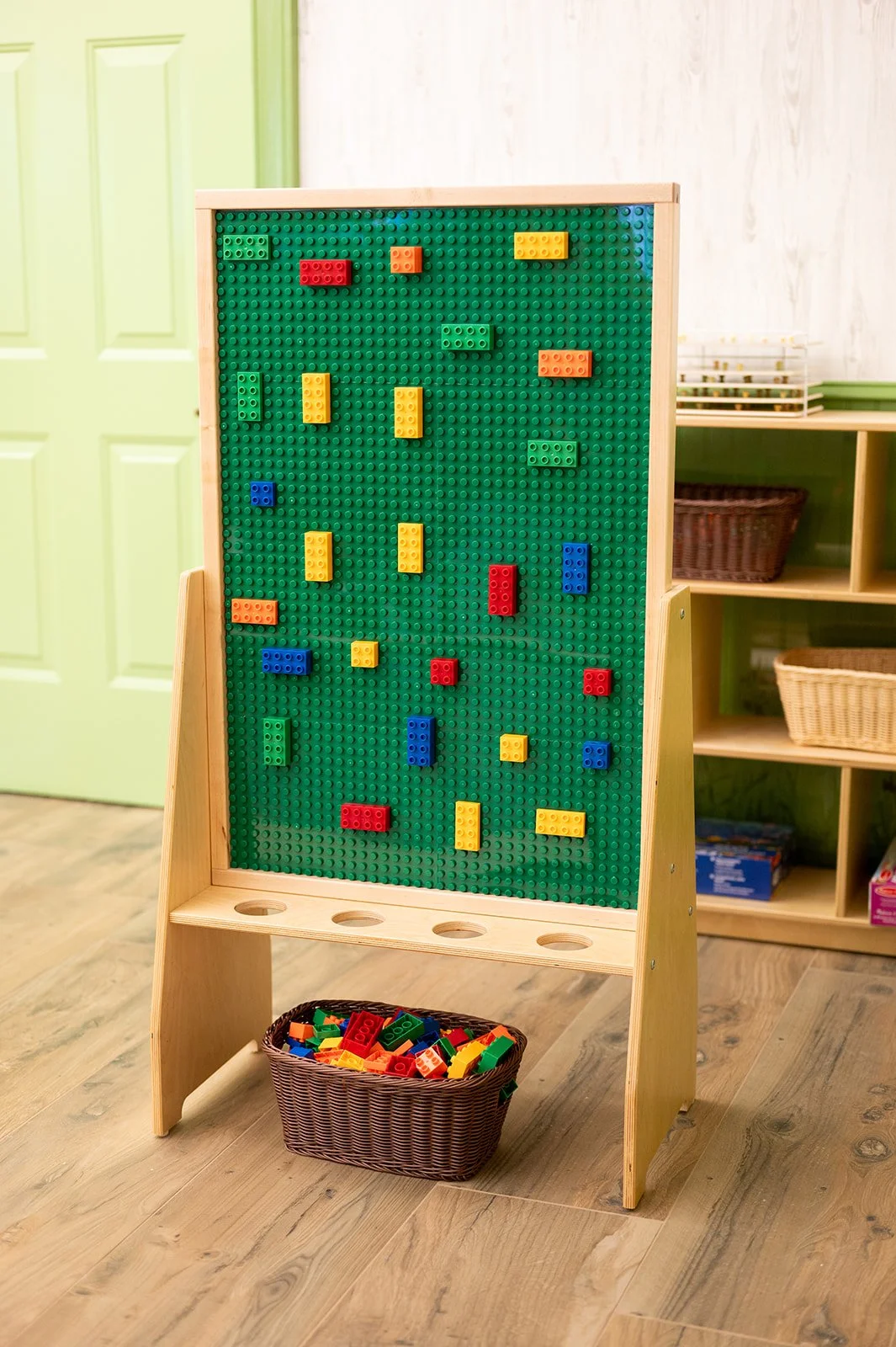 A green pegboard with colorful LEGO bricks attached, standing on a wooden frame with a basket of LEGO pieces underneath, in a room with wooden flooring and light green and white walls.