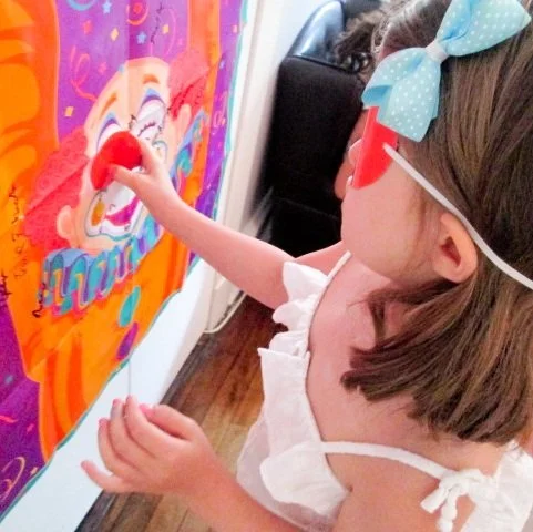 A young girl with a blue polka dot bow, sunglasses, and a white dress is placing a red heart sticker on a colorful, cartoonish wall art.