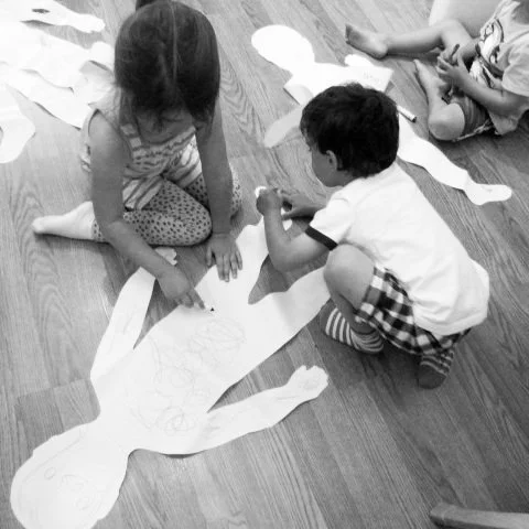 Children cutting out paper shapes and drawing on paper on the floor.