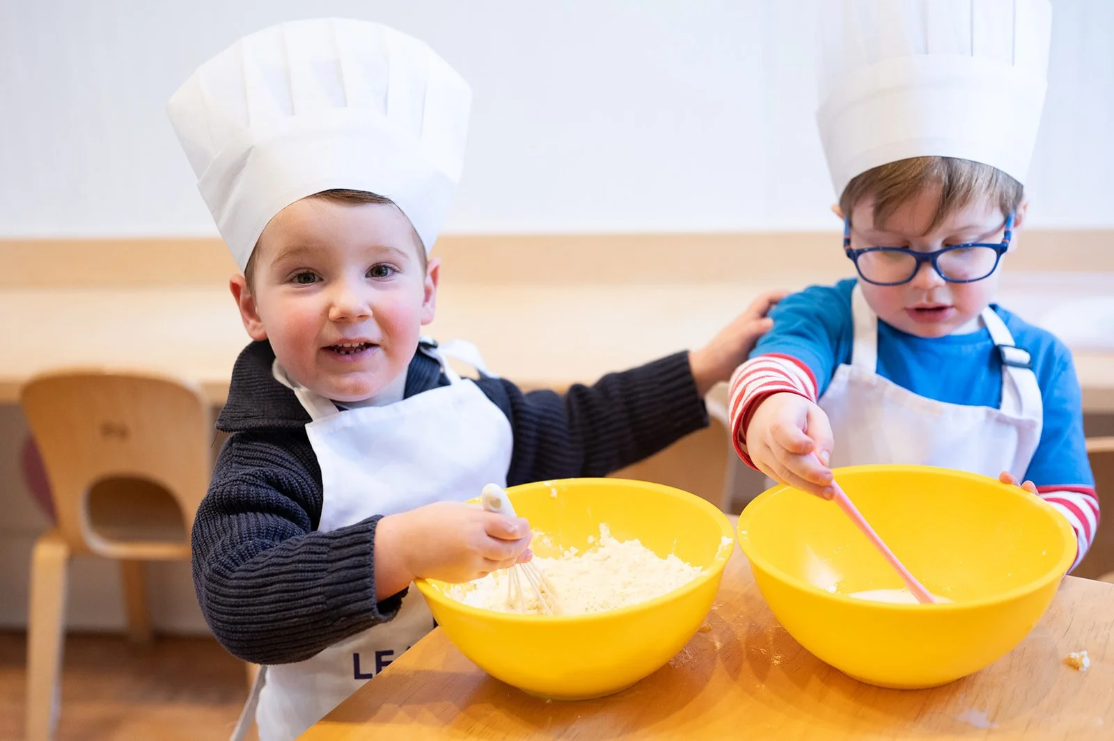 Two young boys wearing chef hats, aprons, and glasses are cooking together at a wooden table. They are mixing ingredients in large yellow bowls.