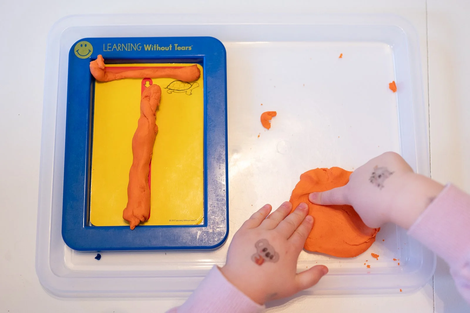 Child playing with orange modeling clay on a white tray, with a blue and yellow learning tool that says "Learning Without Tears" and has a turtle illustration.