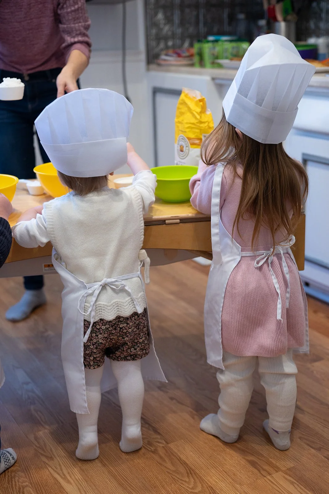 Two children wearing chef hats and aprons, participating in a cooking activity in a kitchen with a table, bowls, and a packet of flour.