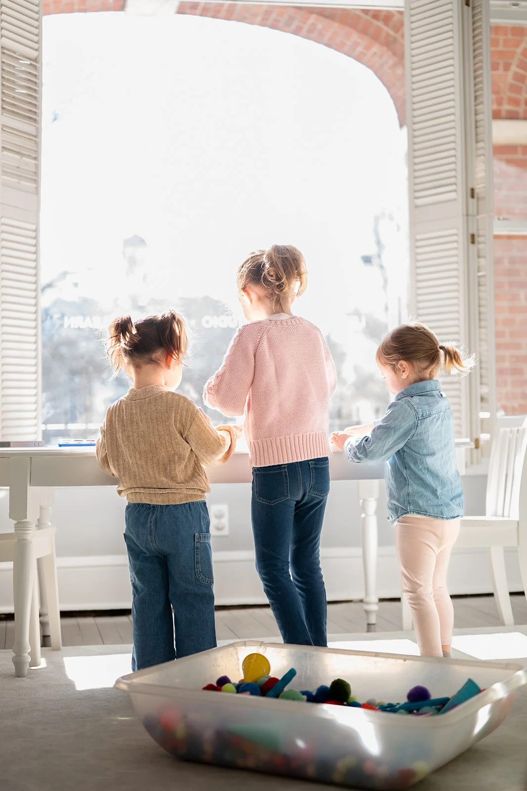 Three children standing by a window, playing with toys on a white table, with a large plastic container filled with colorful toys in the foreground.