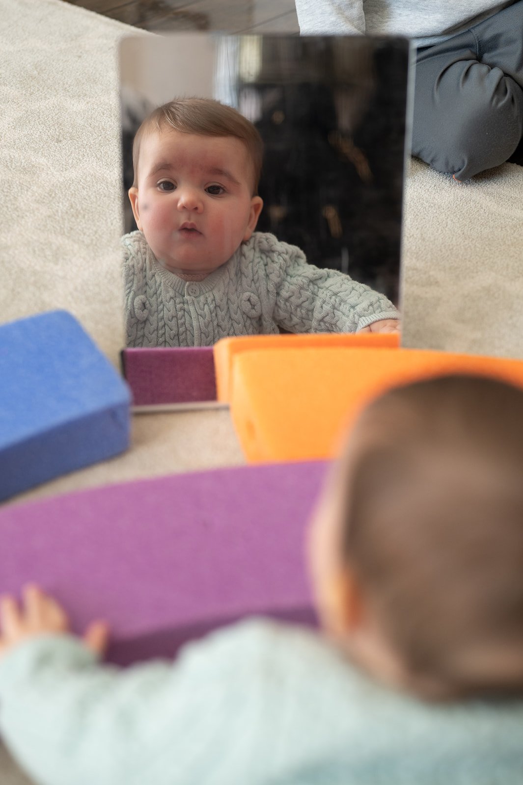 A baby looking into a mirror placed on the floor, with colorful foam blocks in the foreground.