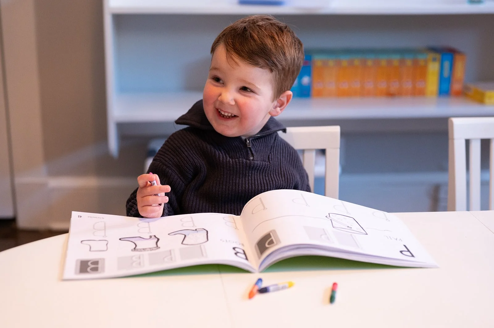 A happy young boy with brown hair sitting at a white table, working on a coloring activity book with a black crayon, with colorful crayons on the table in front of him, in a room with a bookshelf in the background.