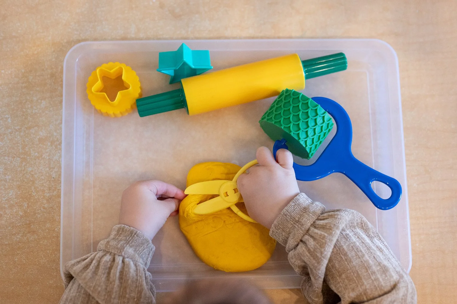 Child's hands playing with yellow and green modeling clay, surrounded by various colorful clay tools and cutters on a clear plastic surface.