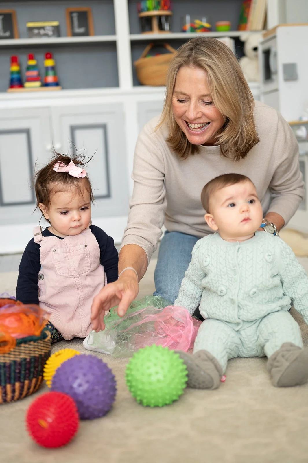 A woman and two young children sitting on the floor playing with textured balls and toys. The woman is smiling and reaching for a green spiky ball. The children, a girl with a pink bow and a boy in a light-colored sweater, are sitting on a carpeted floor in a playroom with shelves and toys in the background.