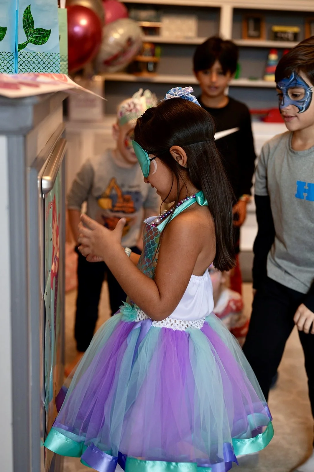 Children dressed as mermaids and superheroes gather around a girl in a mermaid costume, at a birthday party with colorful balloons and decorations.