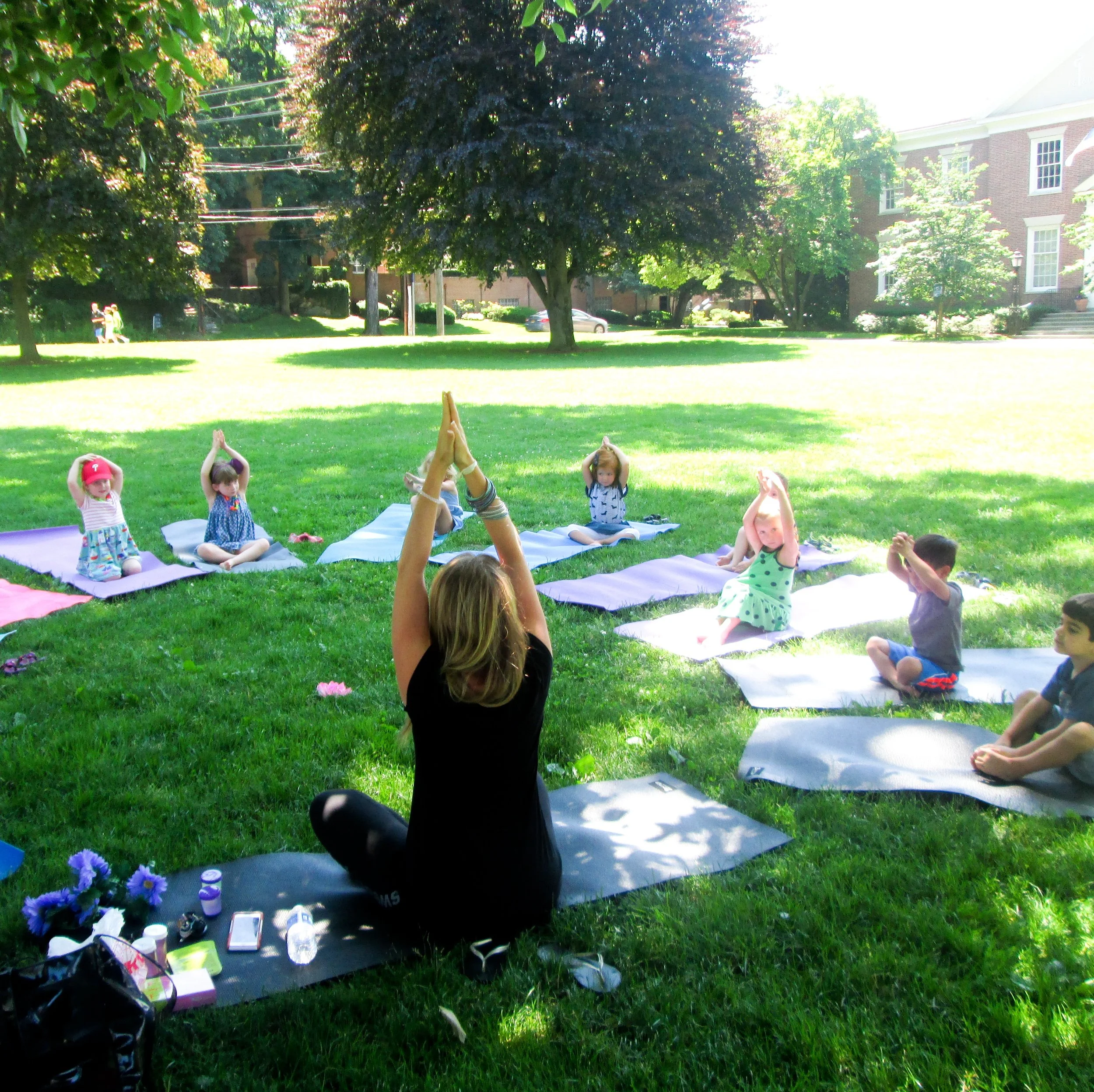 A group of young children and an adult woman practicing yoga outdoors on a grassy park, sitting on yoga mats under the shade of trees, with houses in the background.