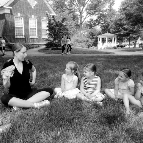 A woman sitting cross-legged on the grass talking to four young girls, all sitting in a row. The woman is holding an ice cream cone, and the girls are listening attentively. In the background, there are two people walking near a gazebo and a brick building.
