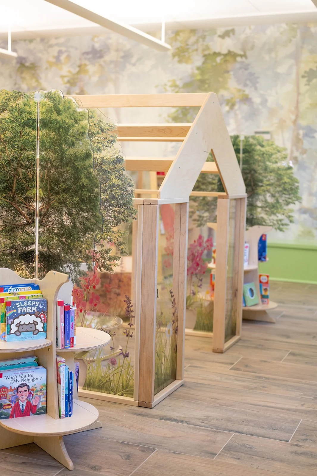 Children's library or play area with wooden house structure, decorated with nature-themed wallpaper, small bookshelf filled with children's books, and a miniature white chair.