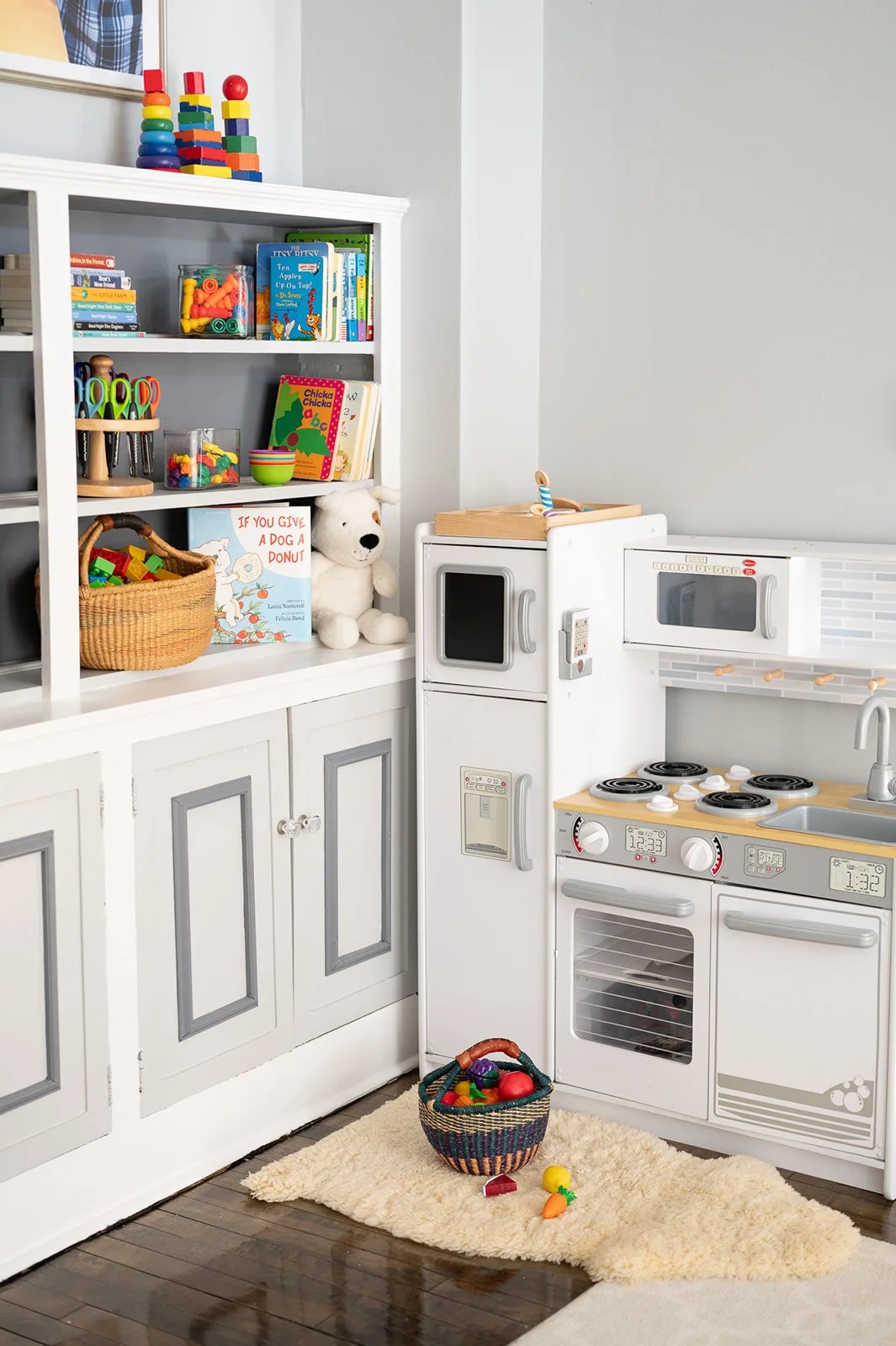 A children's play kitchen setup with toy food, books, and toys including a stuffed animal, with a basket of toys on a beige rug.