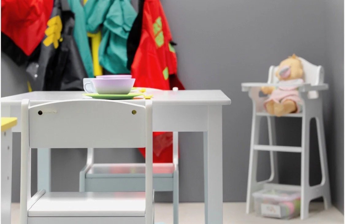 A children's play area with a white table, a purple teacup on a green saucer, and a child's chair. In the background, a high chair holds a teddy bear doll dressed in pink, with a gray wall and colorful jackets hanging on hooks.