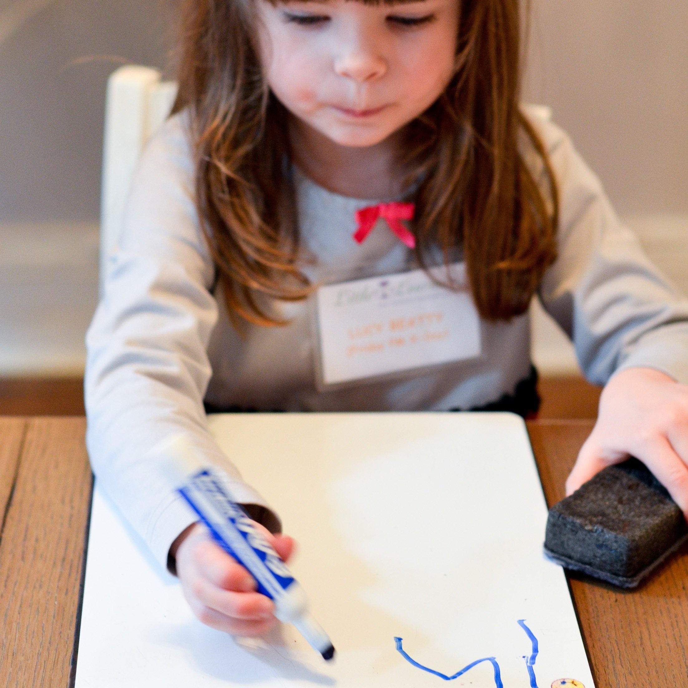 Young girl drawing on a whiteboard with a blue marker, wearing a light-colored shirt with a pink bow, and an identification badge.