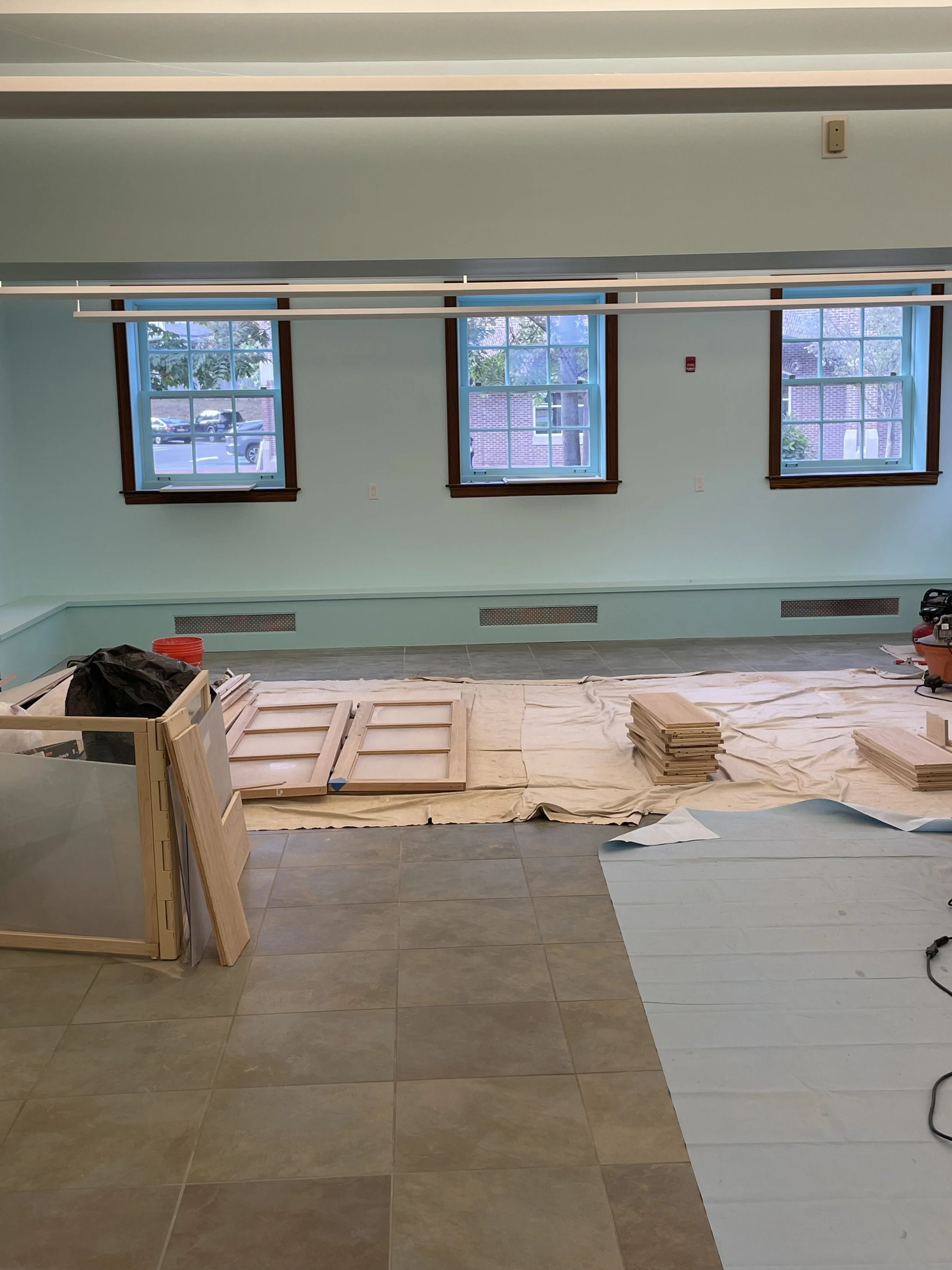 Room under renovation with light green walls, tile flooring, and three windows. Construction materials like wooden panels and tools are laid on the floor.