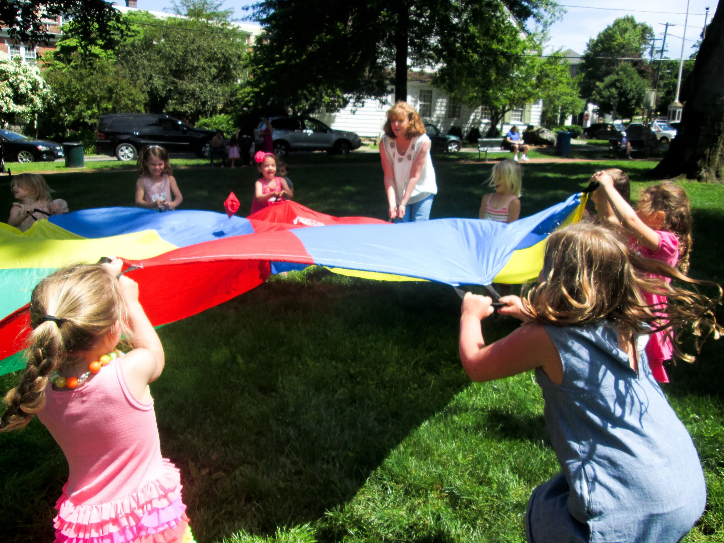 Children and an adult playing with a colorful parachute in a park on a sunny day, with trees, cars, and houses visible in the background.