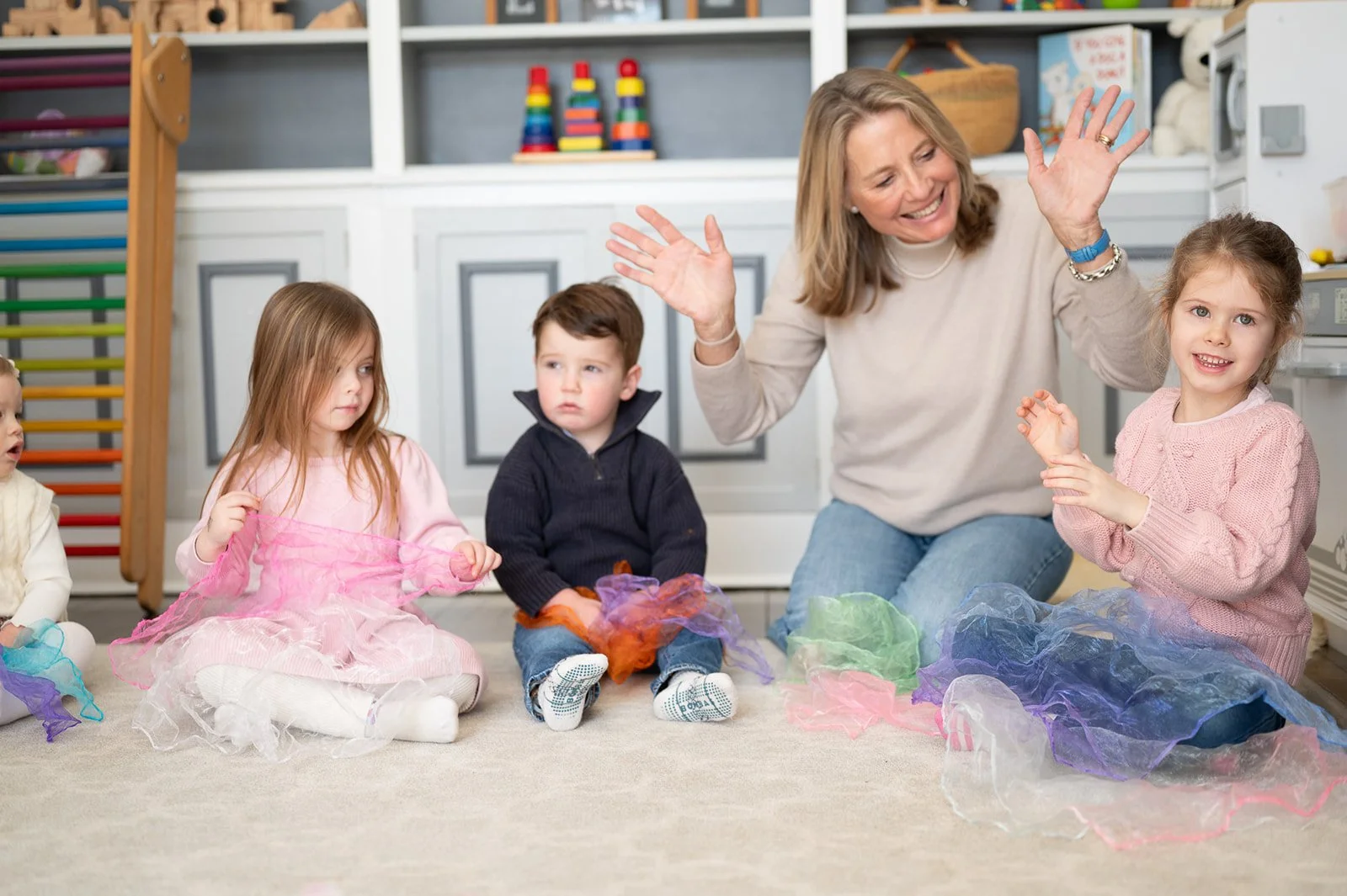 A smiling woman leading a children's activity in a playroom with four kids, sitting on the floor, playing with colorful scarves.