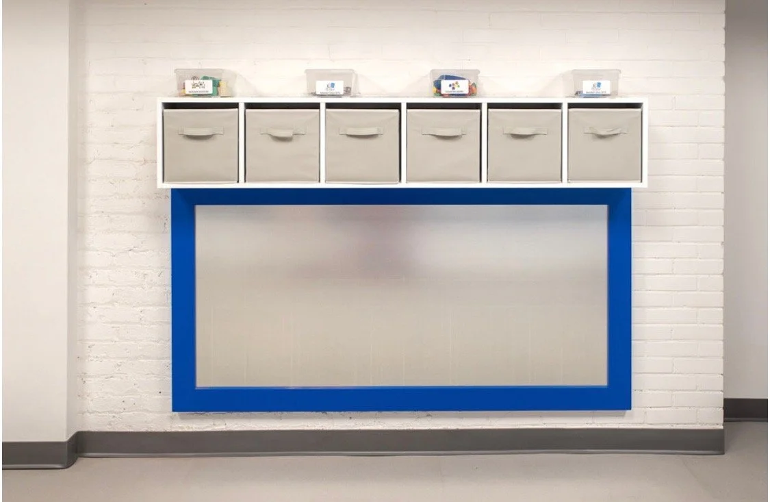 A white brick wall with six small brown and white boxes on a white shelf and a large blue-framed bulletin board.