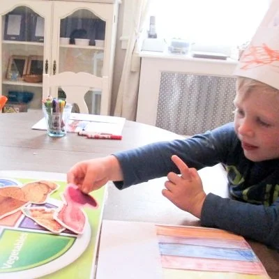 Young boy with a paper crown playing with a dinosaur sticker activity at a table in a bright room.