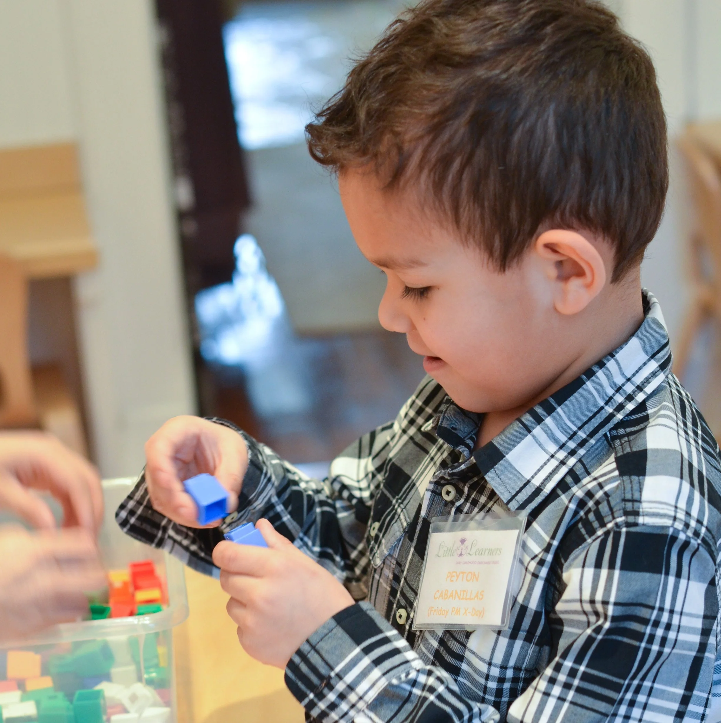 A young boy with brown hair and a checkered shirt playing with colorful building blocks at a table.