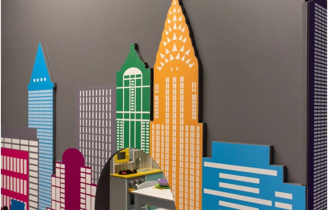 Colorful rooftop cityscape wall art featuring stylized skyscrapers in blue, green, orange, pink, and purple with geometric patterns, next to a small kitchen area with a yellow microwave, purple bowl, and utensils.