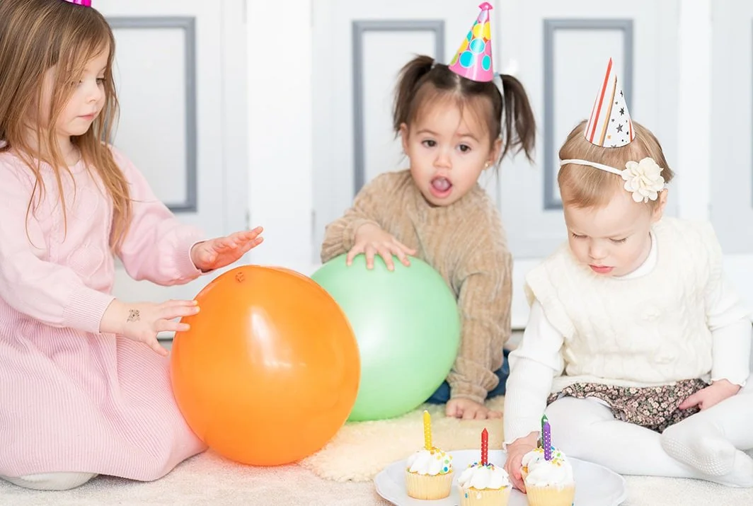 Three young girls celebrating a birthday with party hats, balloons, cupcakes with candles, and a birthday girl sitting on the floor.