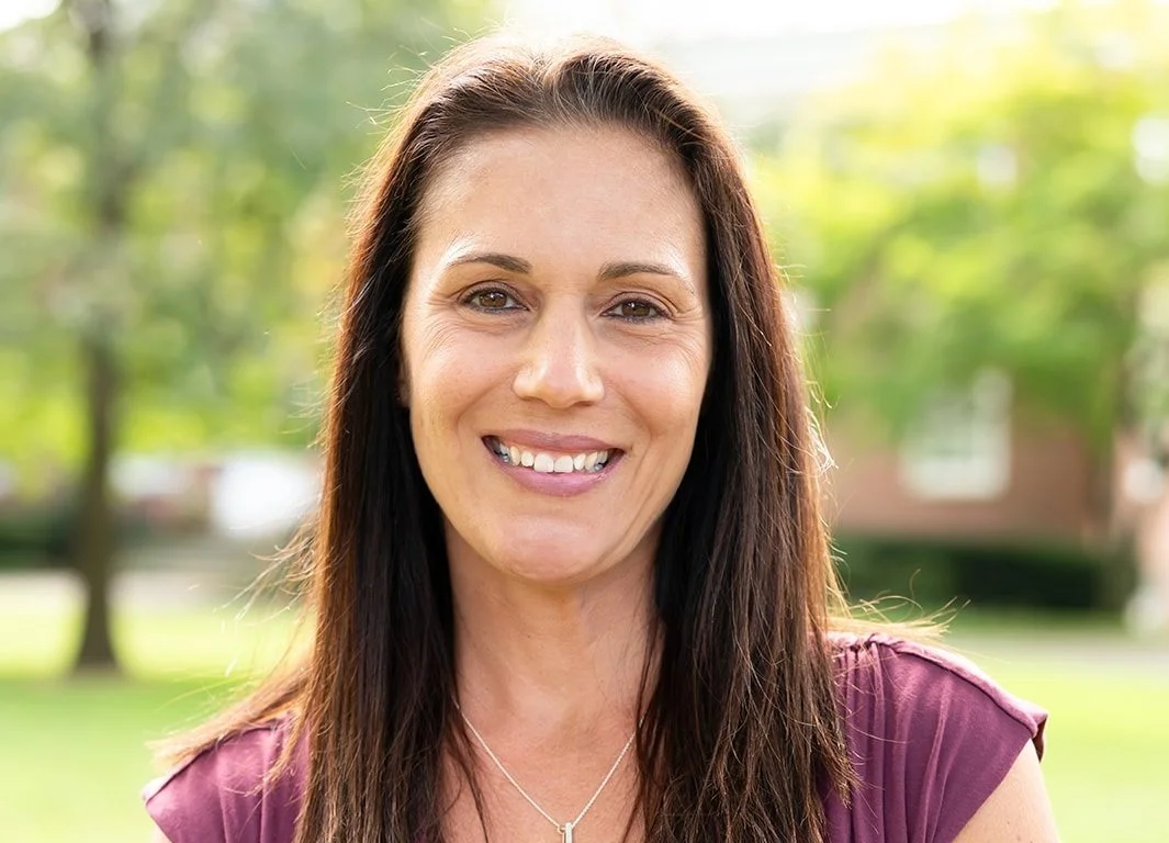 A smiling woman with long dark hair outdoors in a park with green trees in the background.