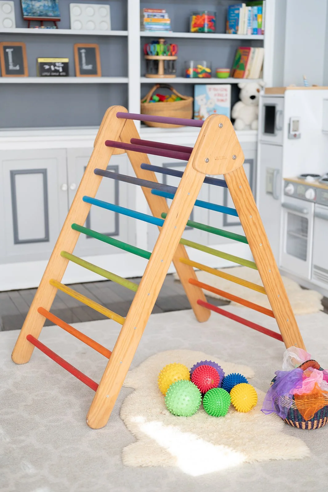 A wooden indoor playground climber with rainbow-colored rungs, placed on a white rug. There are textured balls and a small basket with colorful fabric nearby in a room with bookshelves and kitchen cabinets.