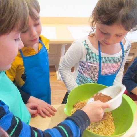 Children wearing aprons gathered around a table, measuring ingredients with a scoop into a large green mixing bowl, preparing to cook or bake.