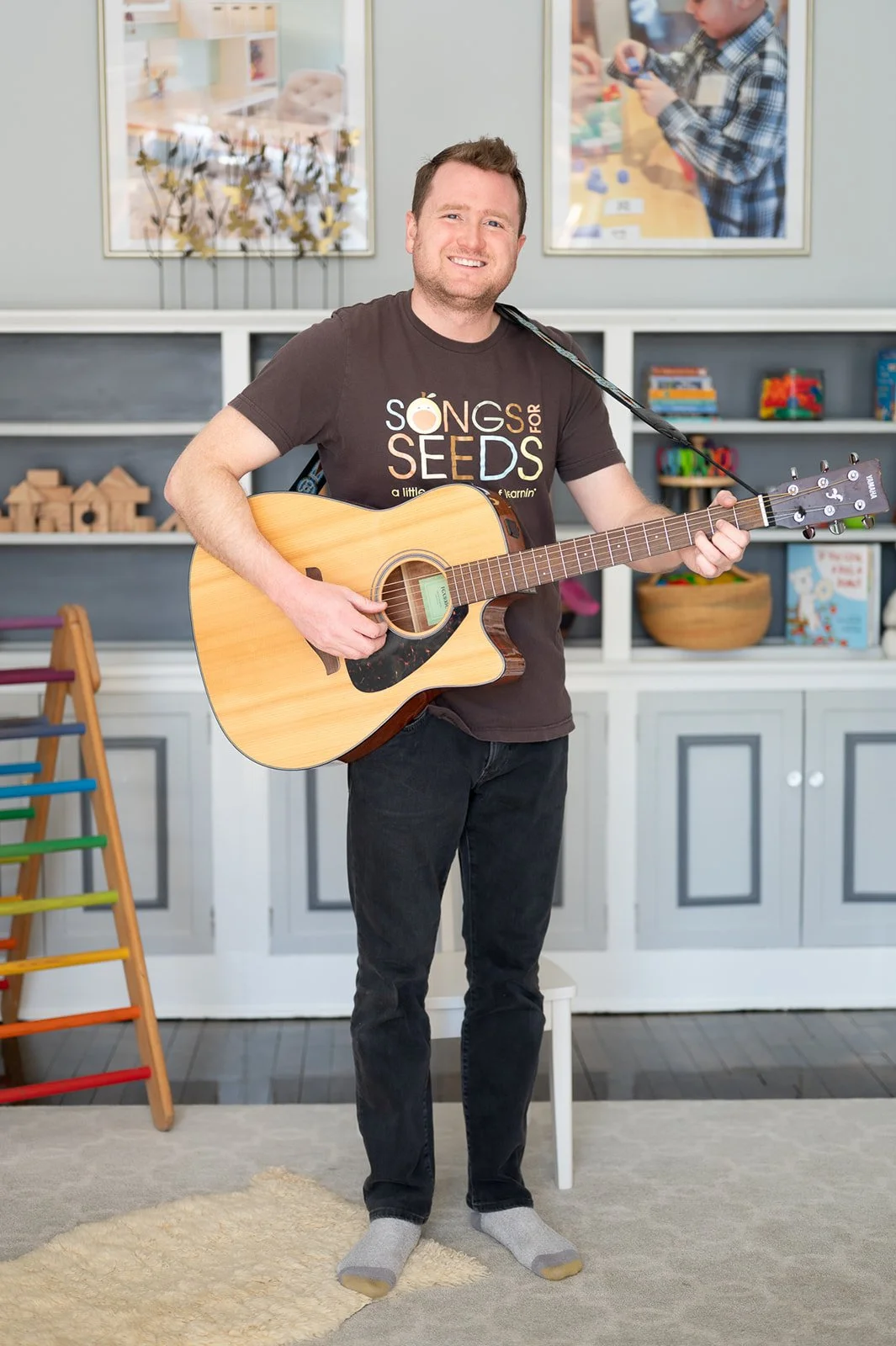 A young man playing an acoustic guitar in a living room or playroom.