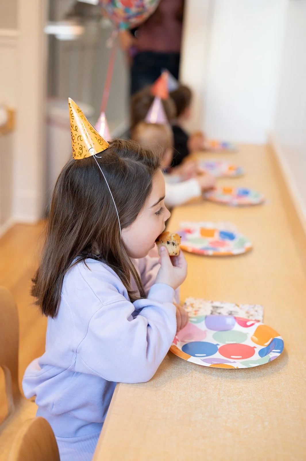 Child in a purple sweatshirt wearing a birthday hat and eating a cupcake at a birthday party, seated at a long table with colorful plates, others children wearing birthday hats in the background.