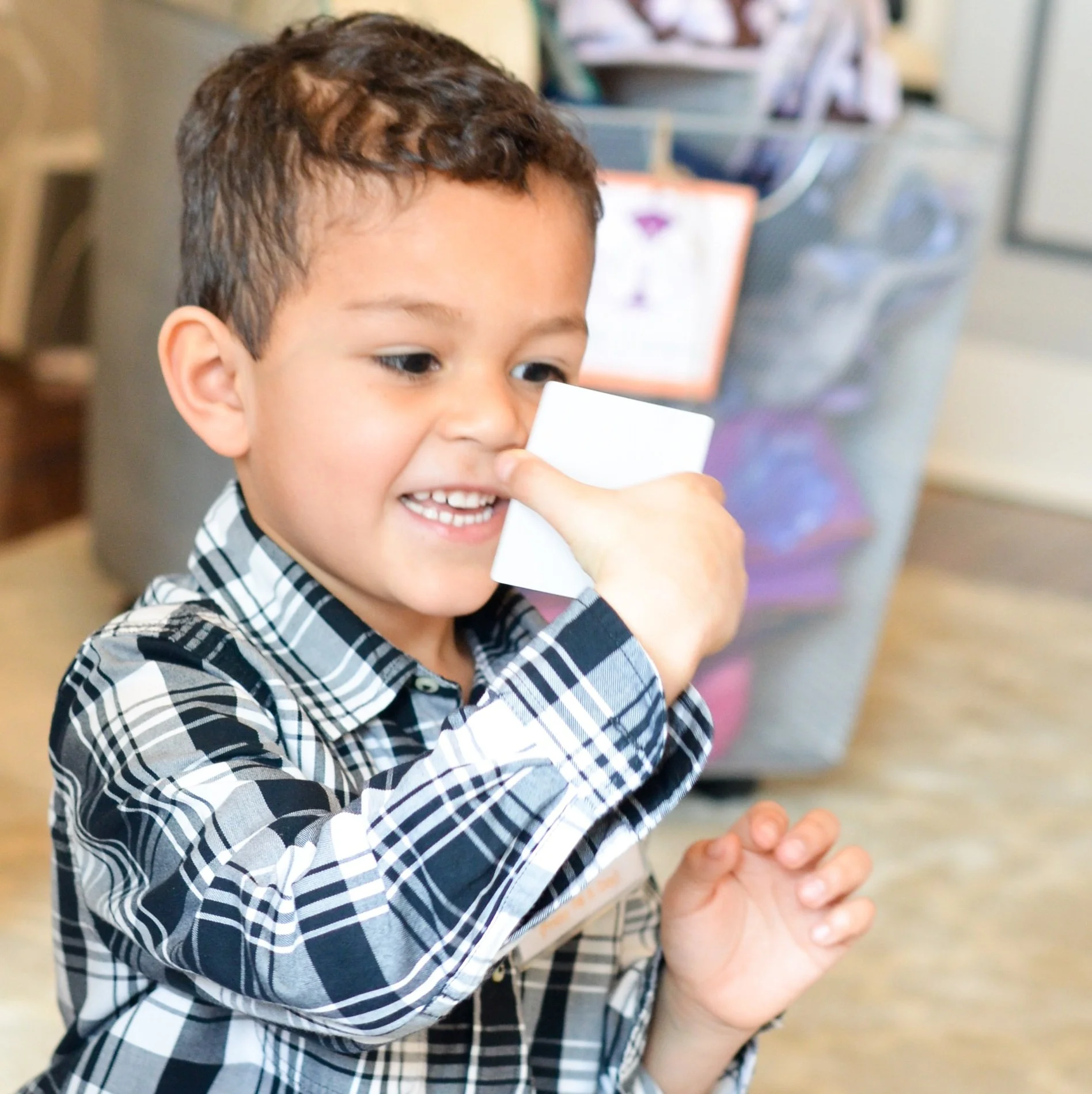 A young boy with dark hair smiling and holding a white card near his nose in a room with a chair and a computer in the background.
