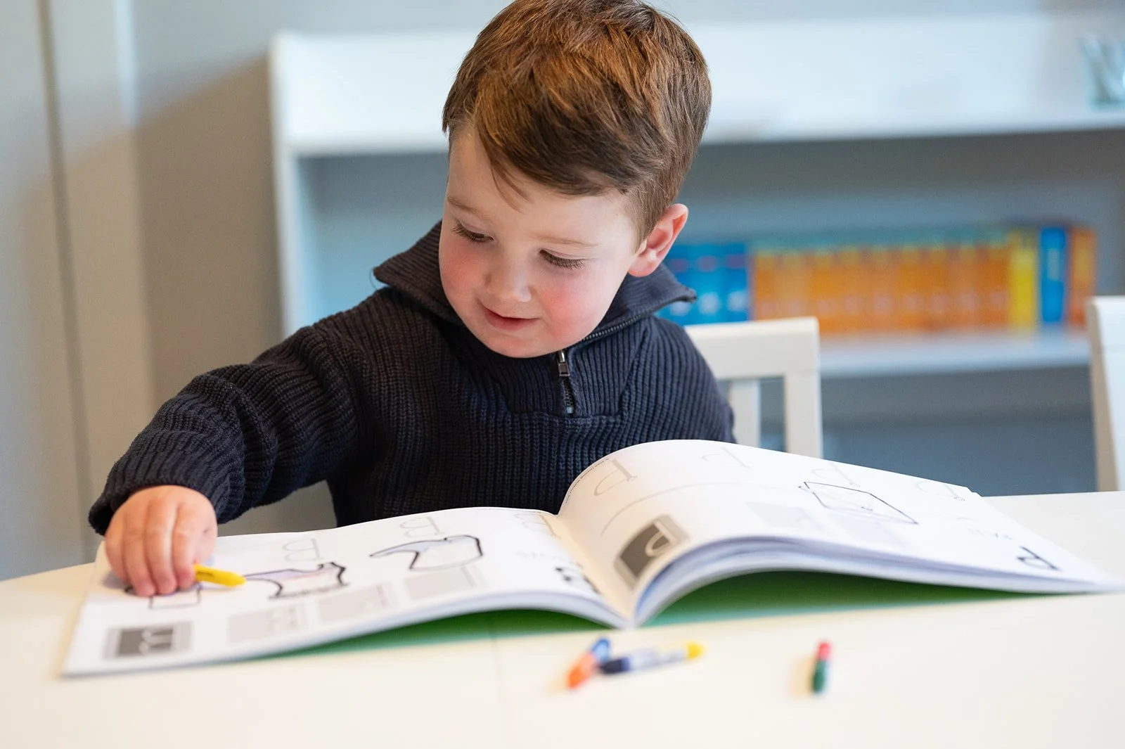 A young boy with brown hair, wearing a black zip-up sweater, sitting at a white table, looking at an open workbook with drawings and words, holding a yellow crayon, with colorful chalks on the table.