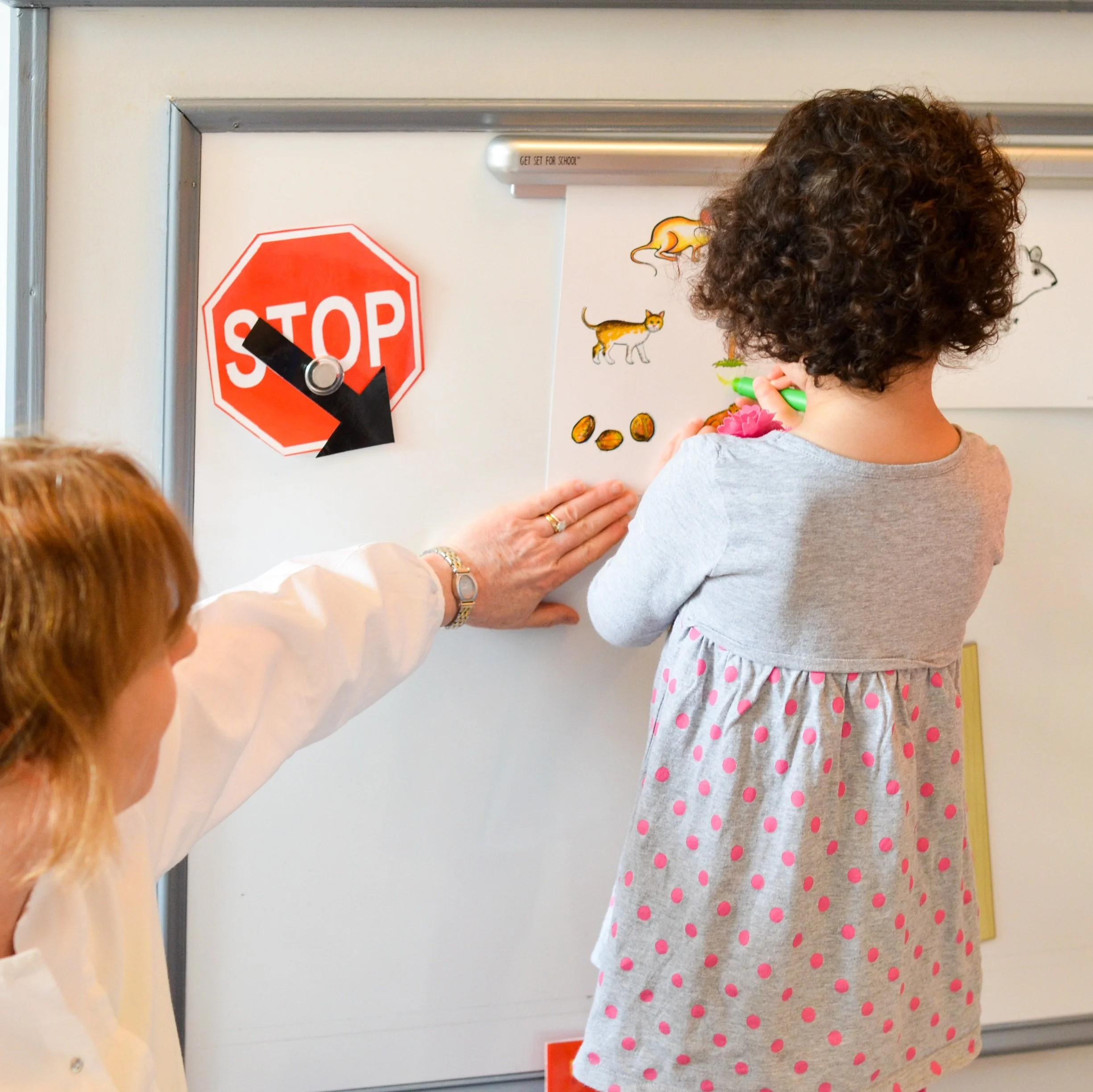 A young girl is coloring pictures of animals and objects on a whiteboard with a green marker, while an adult is pointing to her work.