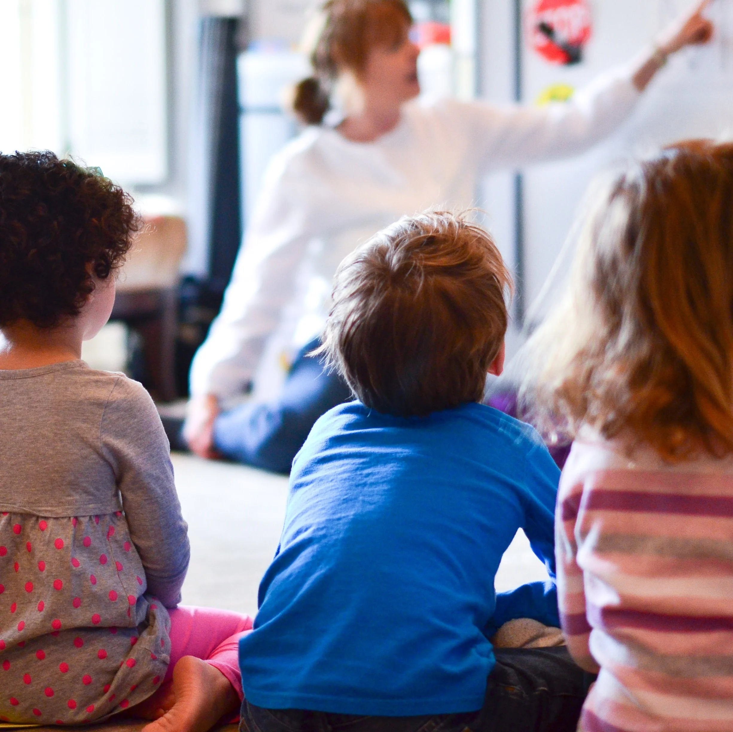 Children sitting on the floor in a classroom, listening to a woman who is teaching and pointing at a whiteboard.