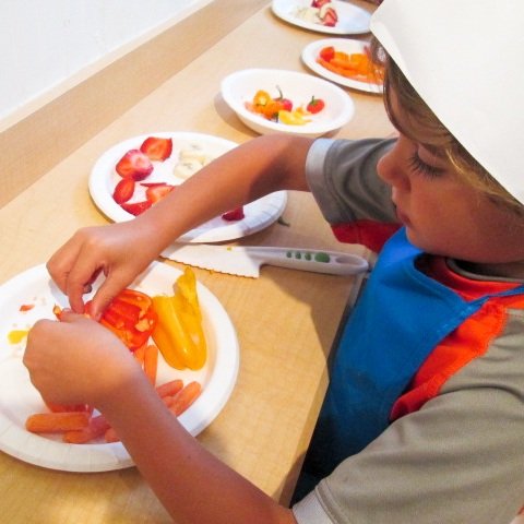 Child preparing fruit platter with sliced strawberries, bananas, and other fruits.