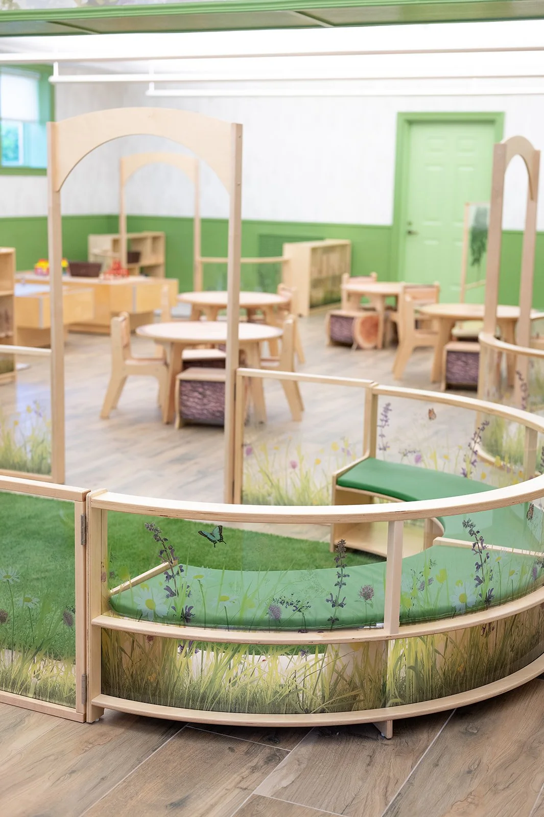An indoor play area for children with wooden furniture, painted green accents, and nature-themed decorations, including glass panels with grass and butterfly illustrations.