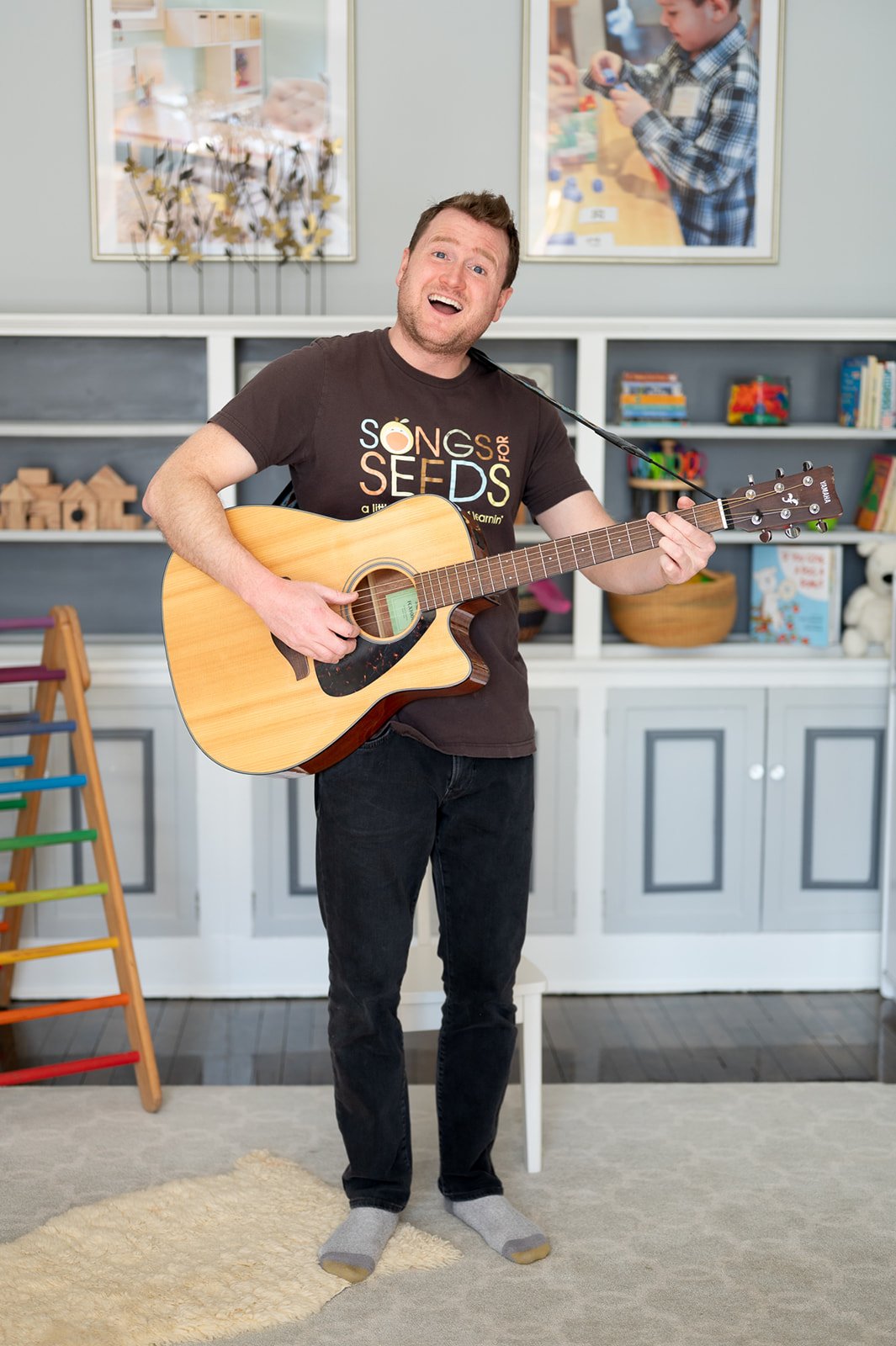 A man with short brown hair and a beard playing an acoustic guitar and singing in a brightly decorated room. He is wearing a black t-shirt with colorful text and black pants, standing on a beige rug. There are children’s toys and artwork on the shelves behind him and a small child in a checkered shirt playing with toys in the background.