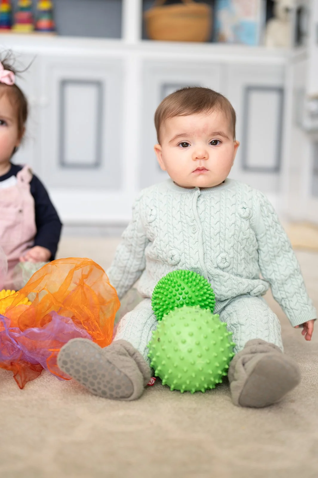 A young child in a light gray sweater and matching pants, sitting on the floor with two green spiky massage balls in front of them. To their left, there is a colorful orange and purple soft textured toy. In the background, another child is partially visible, and the setting appears to be a playroom or living area with white cabinetry.