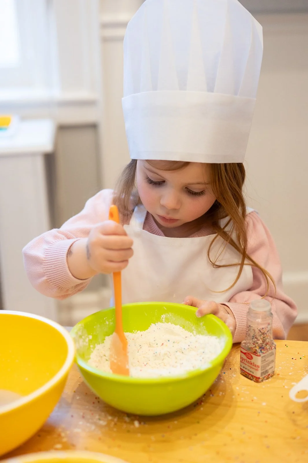A young girl wearing a chef's hat and apron is mixing colorful sprinkles into a bowl of white frosting or dough, with sprinkles scattered on the table.